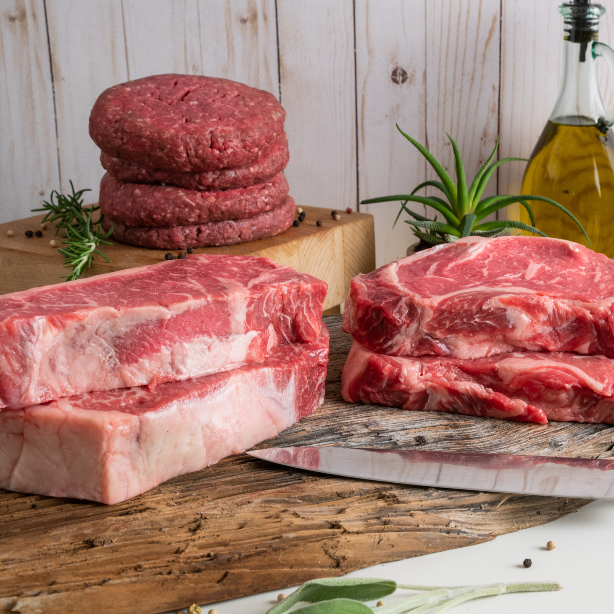 Raw beef cuts and stacked hamburger patties displayed on a rustic wooden cutting board with a knife, herbs, and a bottle of olive oil against a light wood plank background.