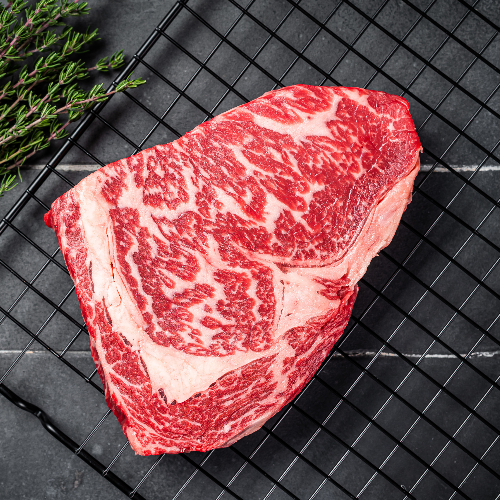 A raw, thickly cut 100% Fullblood American Wagyu ribeye steak resting on a black wire cooling tray, showcasing elite Japanese bloodline intramuscular snowflake marbling at Whole Cow.