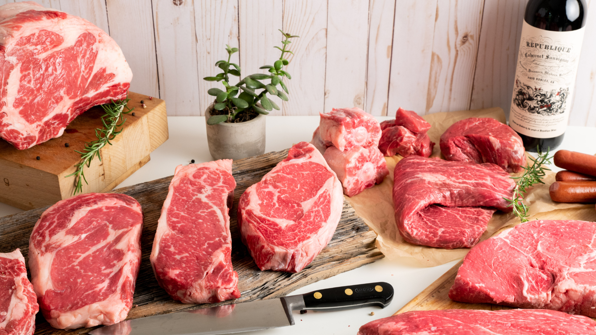A wide 16:9 cinematic selection of raw, custom-cut USDA Prime Black Angus steaks, roasts, and specialty butcher cuts on a rustic board, representing Whole Cow's in-house processing.