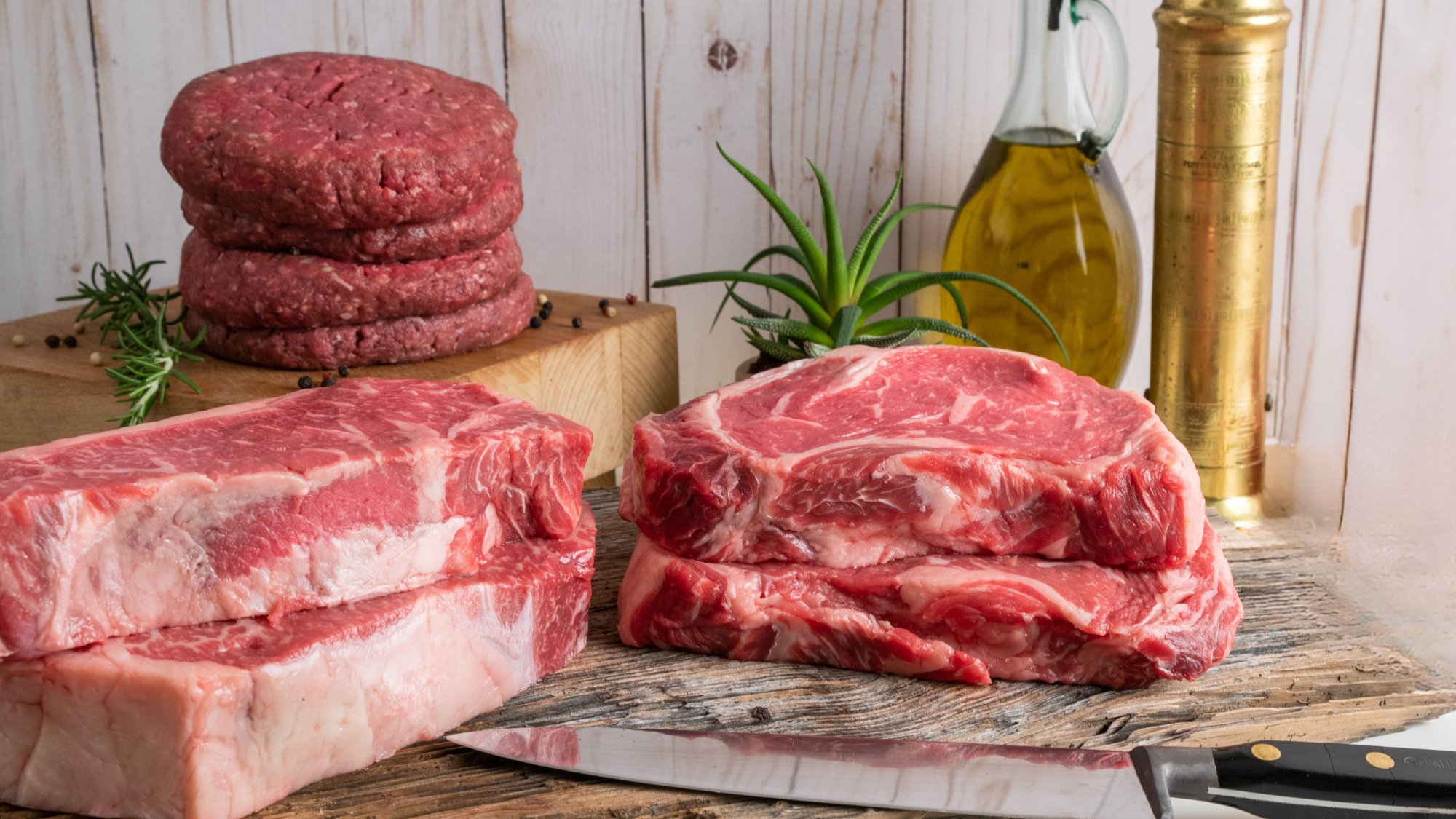 Raw beef USDA Prime steaks and ground beef patties arranged on a rustic wooden cutting board with a knife, olive oil bottle, and brass pepper mill against a white wood plank background.