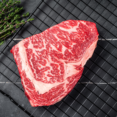 A raw, thickly cut 100% Fullblood American Wagyu ribeye steak resting on a black wire cooling tray, showcasing elite Japanese bloodline intramuscular snowflake marbling at Whole Cow.