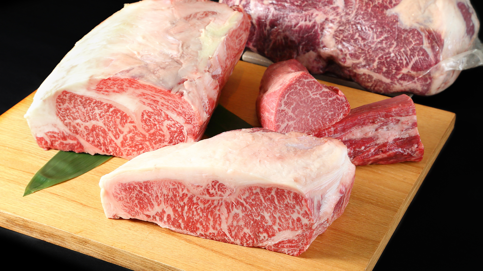 An assortment of raw, uncut 100% Fullblood American Wagyu ribeye and New York strip primal loins arranged on a wooden board, showcasing elite Japanese bloodline snowflake marbling at Whole Cow.
