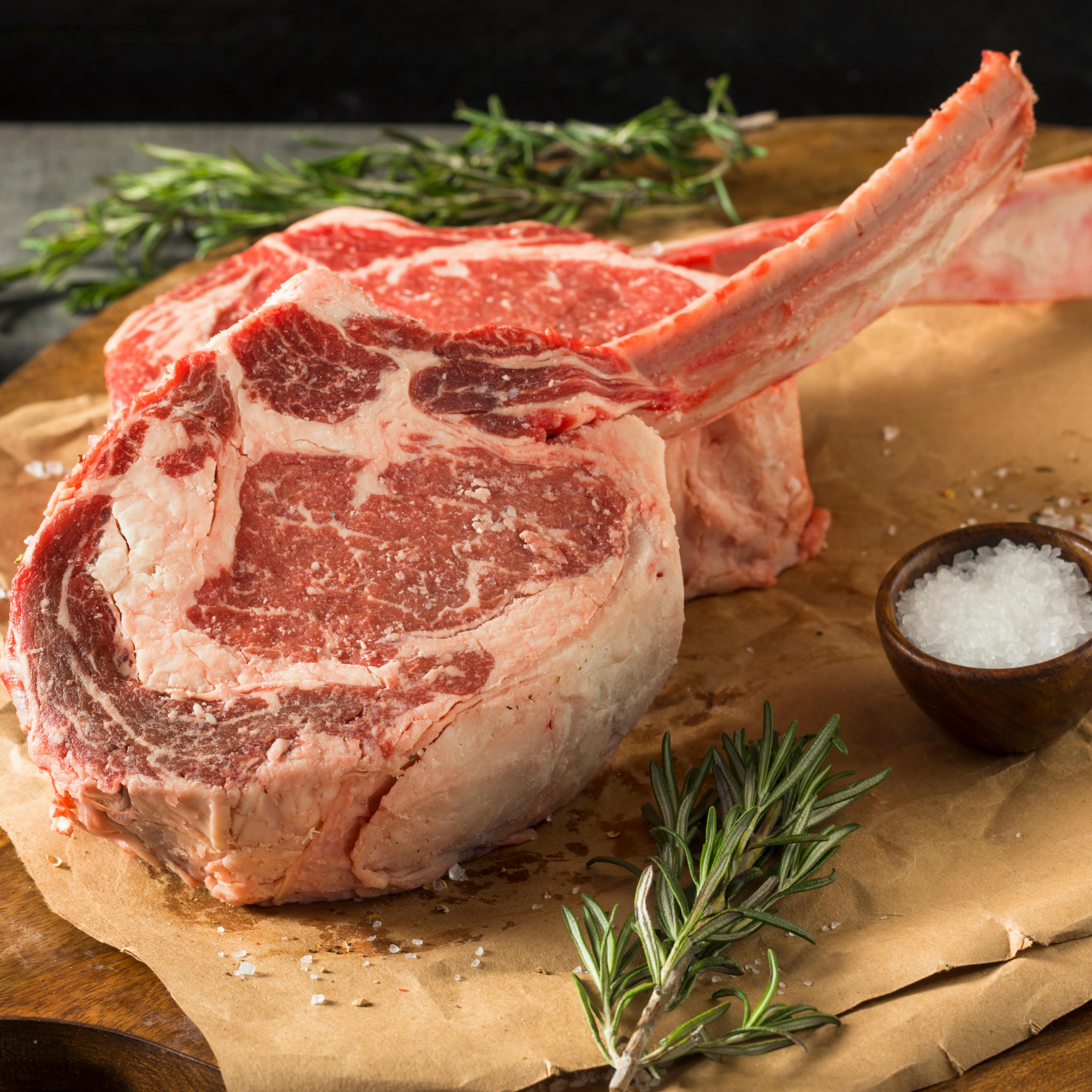 Two raw, thick-cut USDA Prime Black Angus bone-in ribeye steaks seasoned with sea salt and fresh rosemary on butcher paper, processed direct by Whole Cow.