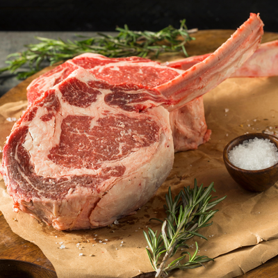 Two raw, thick-cut USDA Prime Black Angus bone-in ribeye steaks seasoned with sea salt and fresh rosemary on butcher paper, processed direct by Whole Cow.